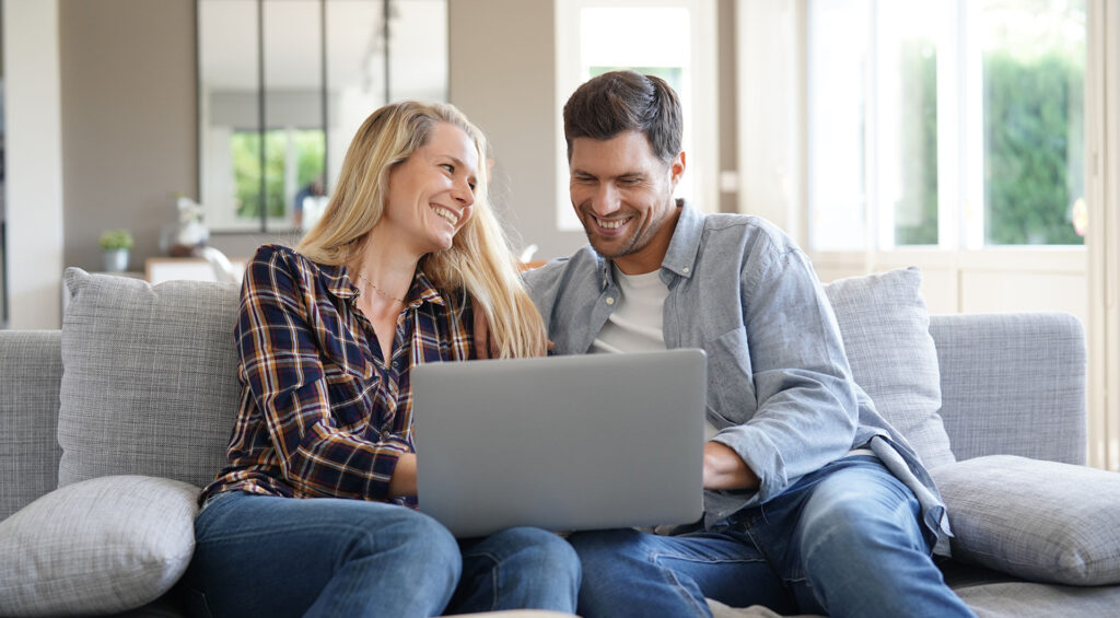Smiling couple sitting together on a sofa reviewing information on a laptop in a bright living room.