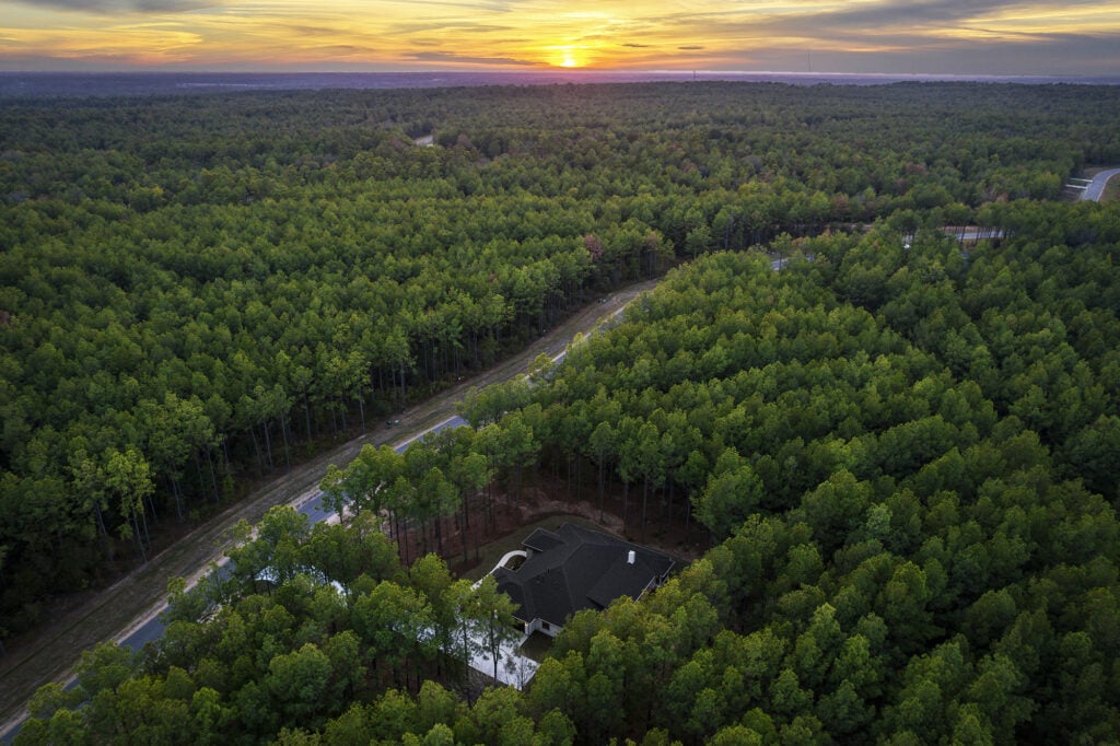 Aerial drone photo of custom home nestled in dense pine forest at sunset with winding driveway and orange glowing sky
