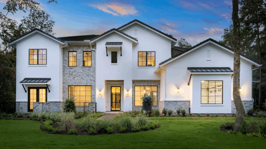 Exterior dusk photo of refined custom home in Carlton Woods Creekside: white brick and stucco with dark metal roof, large black-framed windows, covered porch with warm lighting, landscaped yard, mature trees, and sunset sky in Montgomery County or Greater Houston area