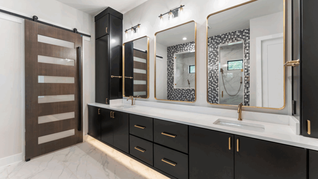 Master bathroom double vanity in a Design Tech Homes custom home: white marble-look countertop with double undermount sinks, gold brass faucets, large backlit black-framed mirror, black sconce lights, marble tiled walls, and sliding barn door in Montgomery County or Greater Houston area