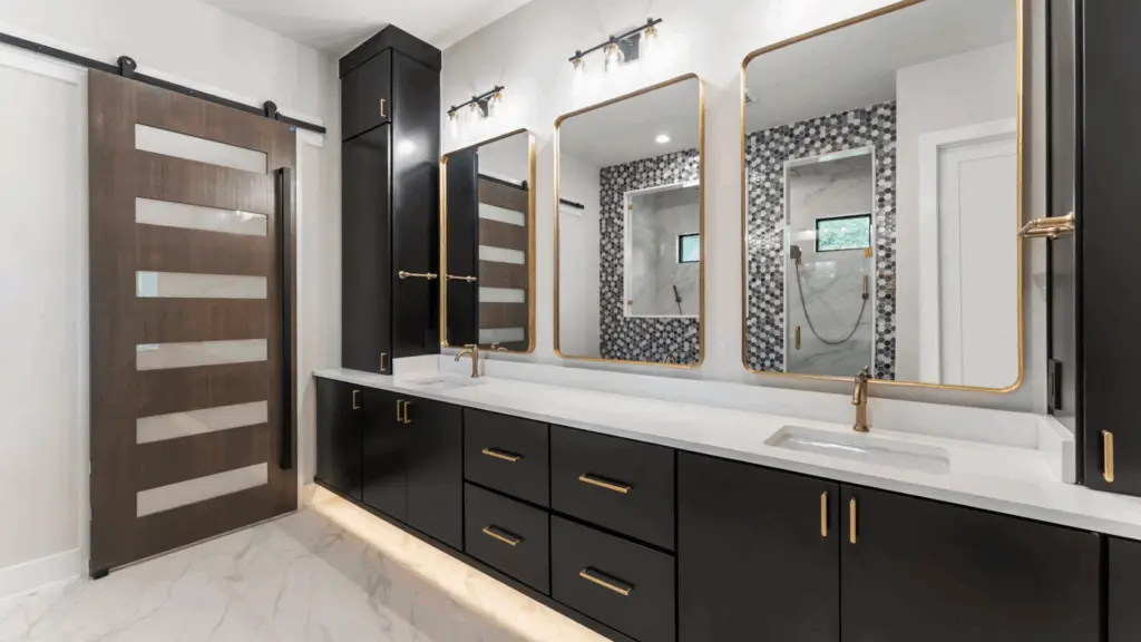 Master bathroom double vanity in a Design Tech Homes custom home: white marble-look countertop with double undermount sinks, gold brass faucets, large backlit black-framed mirror, black sconce lights, marble tiled walls, and sliding barn door in Montgomery County or Greater Houston area