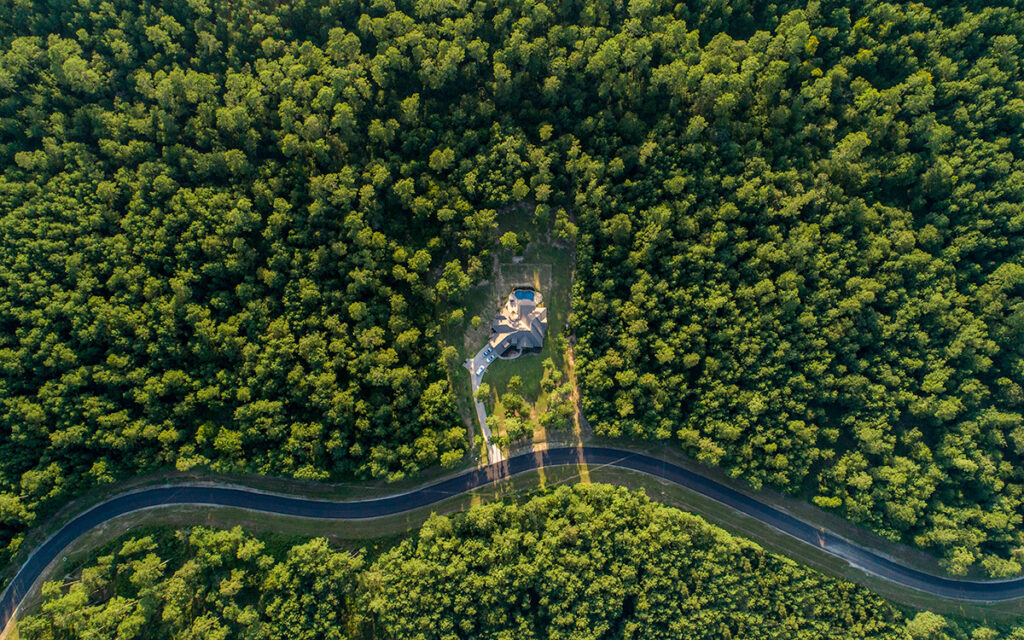 Aerial view of a custom home surrounded by dense forest with winding road access
