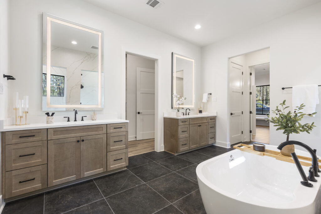 Spacious modern bathroom with double vanities, illuminated mirrors, freestanding soaking tub, and dark tile flooring.