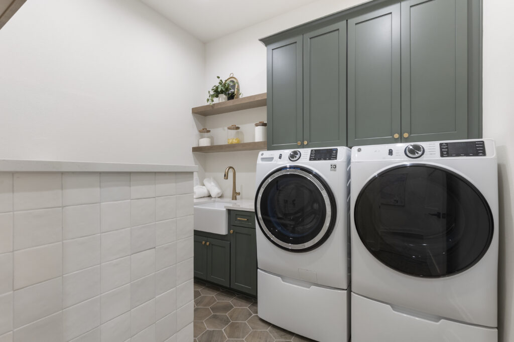 Modern laundry room with front-load washer and dryer, green cabinetry, farmhouse sink, and floating wood shelves.